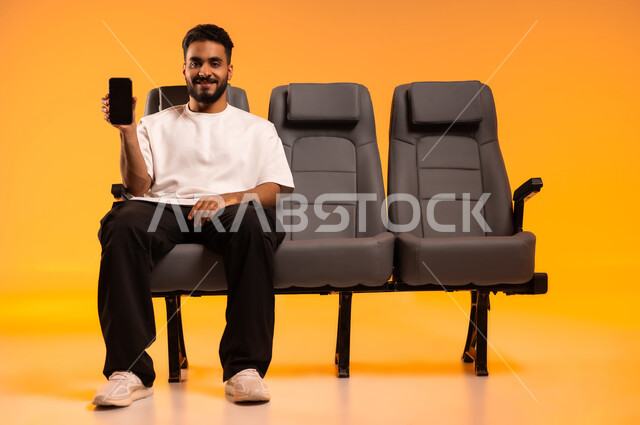 عرض شاشة سوداء فارغة عبر الجوال، استخدام الأجهزة التكنولوجية الحديثة، الاستعداد للسفر في إجازة، الجلوس في صالة المطار, بورتريه لرجل عربي خليجي سعودي يرتدي الزي الكاجوال يجلس على المقعد يقوم باستخدام الهاتف المحمول, أهمية السياحة في السعودية, خلفية برتقالية