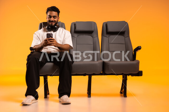 استخدام الأجهزة التكنولوجية الحديثة، الاستعداد للسفر في إجازة، الجلوس في صالة المطار, بورتريه لرجل عربي خليجي سعودي يرتدي الزي الكاجوال يجلس على المقعد يقوم باستخدام الهاتف المحمول, أهمية السياحة في السعودية, رحلة عمل جوية، خلفية برتقالية