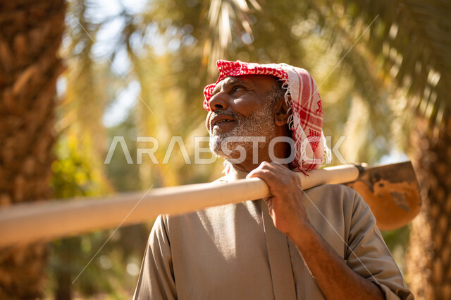 قضاء وقت العمل في الهواء الطلق، الحراثة والاهتمام بالأشجار في مزارع المملكة، استخدام أدوات شعبية قديمة في العمل في المزرعة، فلاح كبير السن عربي خليجي سعودي مبتسم يرتدي الزي الشعبي يسند المجرفة على كتفه وينظر إلى الأعلى في أحد مزارع المملكة العربية السعودية