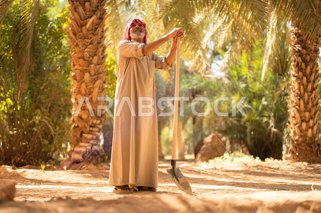 مهن زراعية قديمة محلية، مفهوم مهنة الفلاحة والزراعة، دور الفلاحين السعوديين في المحافظة على التراث الزراعي والإنتاج المحلي، فلاح كبير السن عربي خليجي سعودي يرتدي الزي الشعبي التقليدي ويضع الشماغ على رأسه ينظر إلى الأعلى ويستند على أداة الحفر يستعد لحراثة التربة