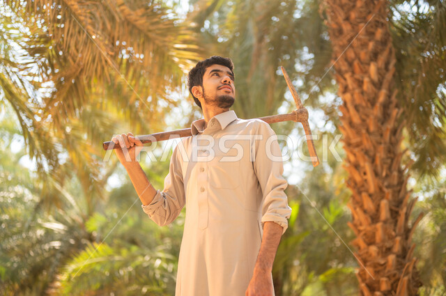 إنهاء العمل بحراثة الأرض في مزرعة النخيل باستخدام أدوات قديمة، فلاح عربي خليجي سعودي يرتدي الزي الشعبي التقليدي يقوم بوضع أداة الحفر على كتفه والنظر إلى الأعلى في مزارع المملكة العربية السعودية بايماءات السعادة والسرور، الاهتمام بتربة المزرعة الغنية والخصبة