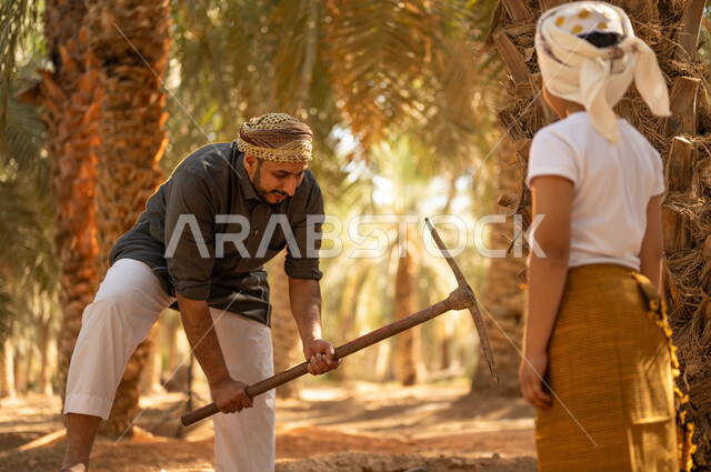 اتقان مهنة الفلاحة والزراعة، مفهوم العائلة السعيدة، تعليم الأبناء على المهن الشعبية، فلاح عربي خليجي سعودي يرتدي الزي الشعبي يقوم بالحفر وبجانبه ابنه ينظر إليه بإيماءات السرور في أحد مزارع النخيل، المحافظة على التراث الزراعي الغني في المملكة العربية السعودية