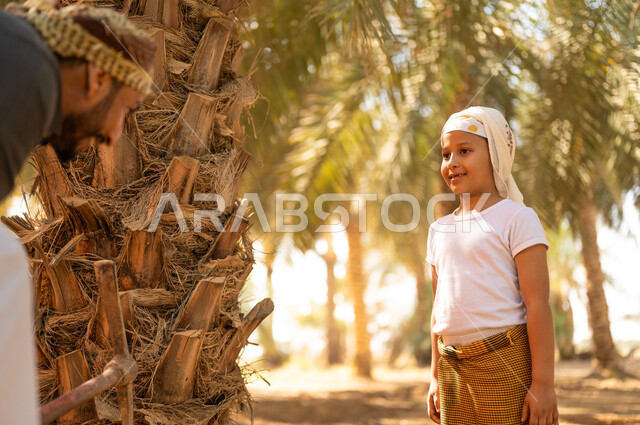 تعليم الأبناء على المهن الشعبية، اتقان مهنة الفلاحة والزراعة، مفهوم العائلة السعيدة، فلاح عربي خليجي سعودي يرتدي الزي الشعبي يقوم بالحفر وبجانبه ابنه ينظر إليه بإيماءات السرور في أحد مزارع النخيل، المحافظة على التراث الزراعي الغني في المملكة العربية السعودية