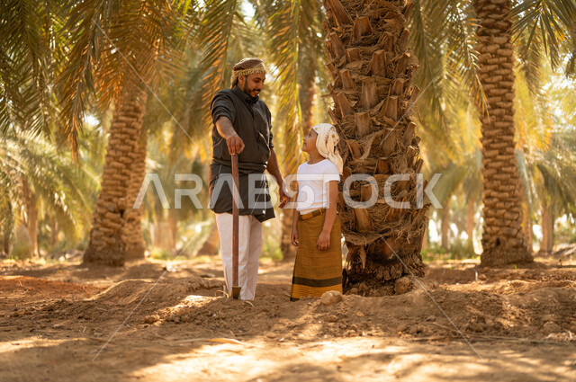 تربية الأبناء على حب المهن الشعبية، اتقان مهنة الفلاحة والزراعة، مفهوم العائلة السعيدة، فلاح عربي خليجي سعودي يرتدي الزي الشعبي يقوم بالوقوف بجانب ابنه وينظر إليه بإيماءات السرور في أحد مزارع النخيل، المحافظة على التراث الزراعي الغني في المملكة العربية السعودية