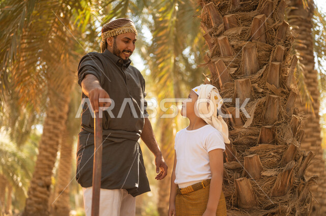 Mastering the profession of farming and agriculture, the concept of a happy family, raising children to love popular professions, a Saudi Arabian Gulf farmer wearing traditional clothing standing next to his son and looking at him with gestures of pleasure in one of the palm farms, preserving the rich agricultural heritage in the Kingdom of Saudi Arabia
