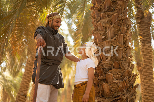 مفهوم العائلة السعيدة، اتقان مهنة الفلاحة والزراعة، تربية الأبناء على حب المهن الشعبية، فلاح عربي خليجي سعودي يرتدي الزي الشعبي يقوم بالوقوف بجانب ابنه وينظر إليه بإيماءات السرور في أحد مزارع النخيل، المحافظة على التراث الزراعي الغني في المملكة العربية السعودية
