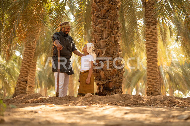 قضاء وقت في الهواء الطلق أثناء العمل في مزارع النخيل المثمرة، مشاركة الأولاد في أعمال الزراعة، فلاح عربي خليجي سعودي مبتسم يرتدي الثوب ويضع الشماغ على رأسه للحماية من الشمس يقف مع ولده أثناء العمل في المزرعة ويمسك بيده أداة الحفر، توريث المهن الزراعية التقليدية