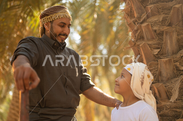 الحفاظ على تراث زراعي غني، الاستعداد لحراثة الأرض الزراعية، صورة مقربة لفلاح عربي خليجي سعودي يرتدي الزي الشعبي التقليدي يحمل بيده أداة الحفر يقف مع ابنه داخل مزارع النخيل المثمرة ينظران إلى الكاميرا بإيماءات السرور، مفهوم المهن الزراعية المحلية