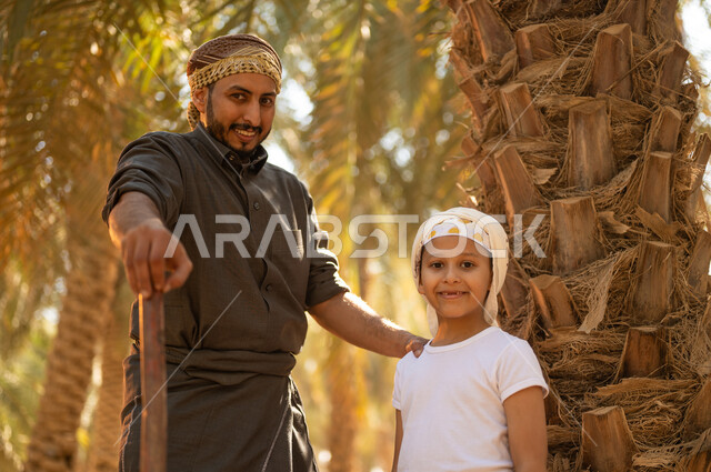 Preserving a rich agricultural heritage, preparing to plow the agricultural land, a close-up photo of a Saudi Arabian Gulf farmer wearing traditional folk costume, holding a drilling tool in his hand, standing with his son inside the fruitful palm farms, with gestures of pleasure, looking at the camera, the concept of the agricultural and farming profession, local agricultural crops