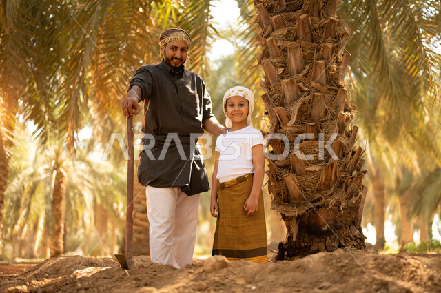 مشاركة الأولاد في أعمال الزراعة، قضاء وقت في الهواء الطلق أثناء العمل في مزارع النخيل المثمرة، فلاح عربي خليجي سعودي مبتسم يرتدي الثوب ويضع الشماغ على رأسه يقف مع ولده أثناء العمل في المزرعة ينظران إلى الكاميرا ويمسك بيده أداة الحفر، توريث المهن الزراعية التقليدية