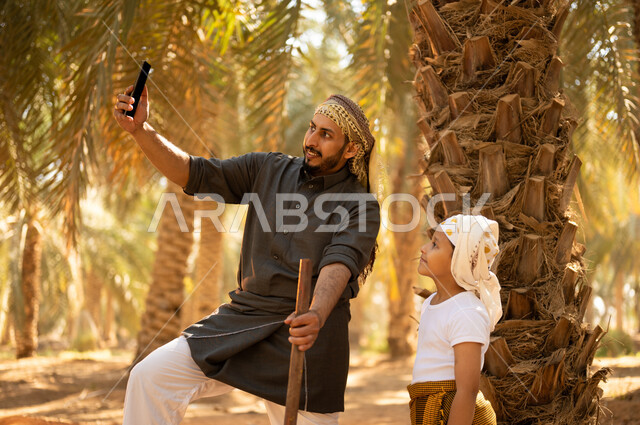Taking memorable selfies while working on fruitful palm farms, children participating in agricultural work, spending time outdoors, a smiling Saudi Arabian Gulf farmer wearing a thobe and wearing a shemagh on his head to protect from the sun, standing with his son while working on the farm, passing on traditional agricultural occupations
