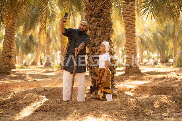 مشاركة الأولاد في أعمال الزراعة، قضاء وقت العمل في الهواء الطلق، فلاح عربي خليجي سعودي مبتسم يرتدي الثوب ويضع الشماغ على رأسه للحماية من الشمس يقف مع ولده أثناء العمل في المزرعة، أخذ صور السيلفي للذكرى أثناء العمل في مزارع النخيل المثمرة، توريث المهن الزراعية التقليدية