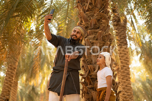 قضاء وقت العمل في الهواء الطلق، فلاح عربي خليجي سعودي مبتسم يرتدي الثوب ويضع الشماغ على رأسه للحماية من الشمس يقف مع ولده أثناء العمل في المزرعة، أخذ صور السيلفي للذكرى أثناء العمل في مزارع النخيل المثمرة، مشاركة الولد في أعمال الزراعة، توريث المهن الزراعية التقليدية