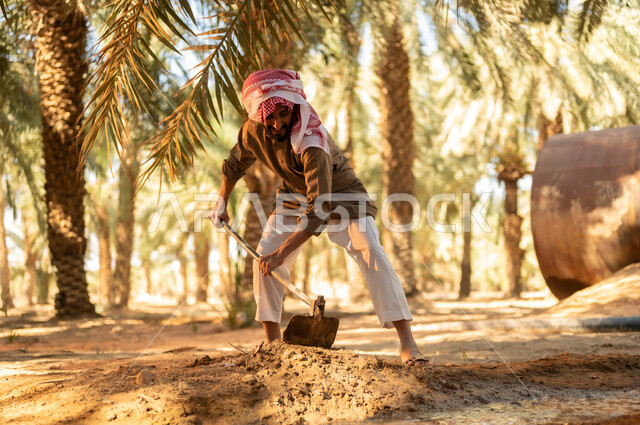 العمل في المزرعة في الري وسقاية الأراضي الزراعية تربة غنية وثمار ذات جودة في المملكة العربية السعودية, زراعة الأشجار المثمرة المحلية, مزارع عربي خليجي سعودي يرتدي الثوب والعمامة يقوم بحفر طريق للماء باستخدام المجرفة ليصل إلى كافة المزروعات، مهن تقليدية محلية