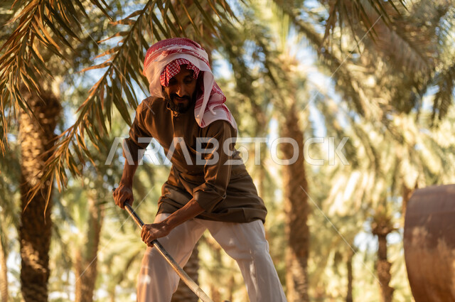 تربة غنية وثمار ذات جودة، زراعة الأشجار المثمرة المحلية, مزارع عربي خليجي سعودي يرتدي الثوب والعمامة يقوم بحفر طريق للماء باستخدام المجرفة ليصل إلى كافة المزروعات، العمل في المزرعة في أعمال الري وسقاية الأراضي الزراعية في المملكة العربية السعودية, مهن تقليدية محلية