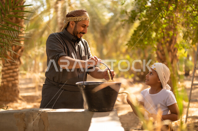 ايماءات السرور والبهجة أثناء العمل داخل مزارع النخيل المثمرة في المملكة، استعمال الطرق القديمة والشعبية في الحصول على الماء، فلاح عربي خليجي سعودي يرتدي الثوب والعمامة يقوم بمساعدة ولده للحصول على المياه من البئر في المزرعة، توريث المهن الزراعية التقليدية المحلية