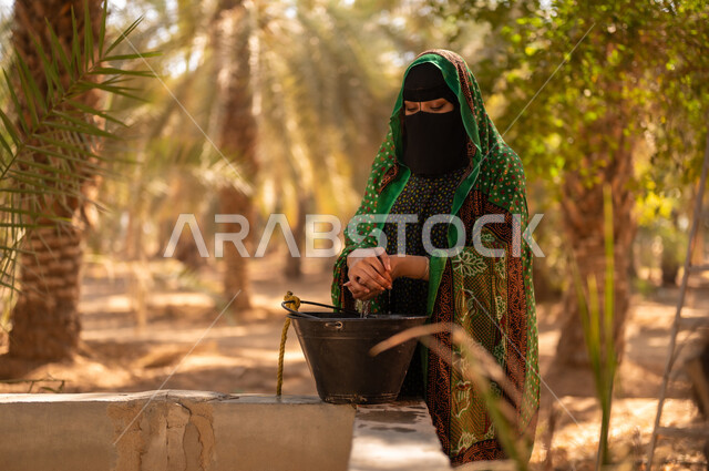 Using old and popular methods to obtain water, women’s participation in work within the fruitful palm farms in the Kingdom, a Saudi Gulf Arab woman wearing traditional folk dress and the burqa washing her hands using well water on the farm, local traditional agricultural occupations