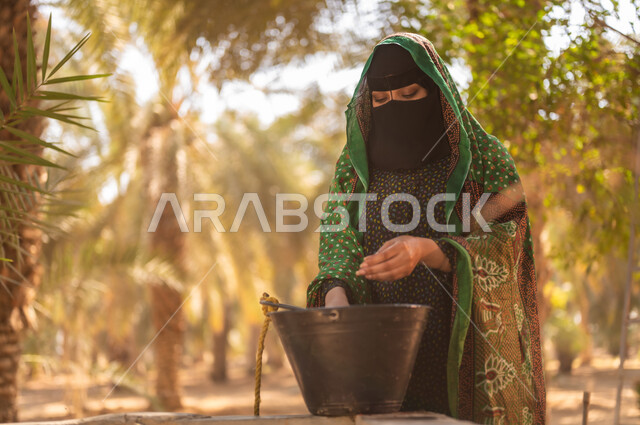 مشاركة المرأة في العمل داخل مزارع النخيل المثمرة في المملكة، استعمال الطرق القديمة والشعبية في الحصول على الماء، إمرأة عربية خليجية سعودية ترتدي الزي الشعبي التقليدي والبرقع تقوم بغسل يديها باستخدام مياه البئر في المزرعة، مهن زراعية تقليدية محلية