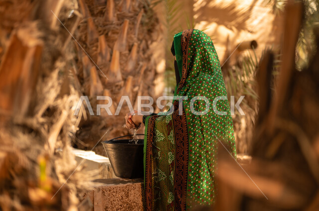 مشاركة المرأة في العمل داخل مزارع النخيل المثمرة في المملكة، استعمال الطرق القديمة والشعبية في الحصول على الماء، إمرأة عربية خليجية سعودية ترتدي الزي الشعبي التقليدي والبرقع تقوم بغسل يديها باستخدام مياه البئر في المزرعة، مهن زراعية تقليدية محلية