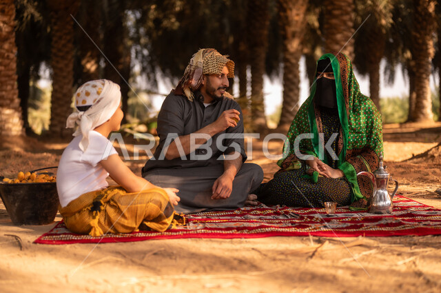 أخذ قسط من الراحة وتبادل الأحاديث مع العائلة في مزارع النخيل المثمرة، قضاء وقت الراحة في الهواء الطلق، فلاح عربي خليجي سعودي مبتسم يرتدي الثوب ويضع الشماغ على رأسه للحماية من الشمس ويجلس على الأرض مع زوجته وولده في المزرعة، مهن زراعية تقليدية