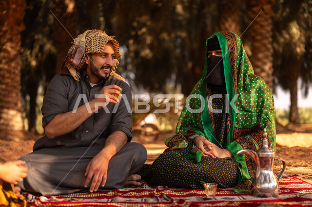 قضاء وقت الراحة في الهواء الطلق، أخذ قسط من الراحة وتبادل الأحاديث في مزارع النخيل المثمرة، فلاح عربي خليجي سعودي مبتسم يرتدي الثوب ويضع الشماغ على رأسه للحماية من الشمس ويجلس على الأرض مع زوجته في المزرعة، مهن زراعية تقليدية، ايماءات السعادة والبهجة