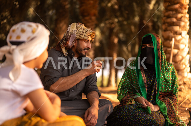 Spending rest time outdoors, taking a break and exchanging conversations with the family in the fruitful palm farms, a smiling Saudi Arabian Gulf farmer wearing a thobe and placing a shemagh on his head to protect from the sun, sitting on the ground with his wife and son on the farm, traditional agricultural occupations