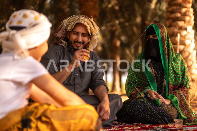 Taking a break and exchanging conversations with the family in the fruitful palm farms, spending rest time in the open air, a smiling Saudi Arabian Gulf farmer wearing a thobe and placing a shemagh on his head to protect from the sun, sitting on the ground with his wife and son on the farm, traditional agricultural occupations
