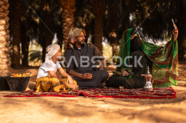 توريث المهن الزراعية التقليدية، قضاء وقت الراحة في الهواء الطلق مع العائلة، فلاح عربي خليجي سعودي مبتسم يرتدي الثوب ويضع الشماغ على رأسه للحماية من الشمس ويجلس على الأرض مع زوجته وولده في المزرعة، النظر إلى الكاميرا وأخذ صور السيلفي للذكرى بعد العمل في مزارع النخيل المثمرة