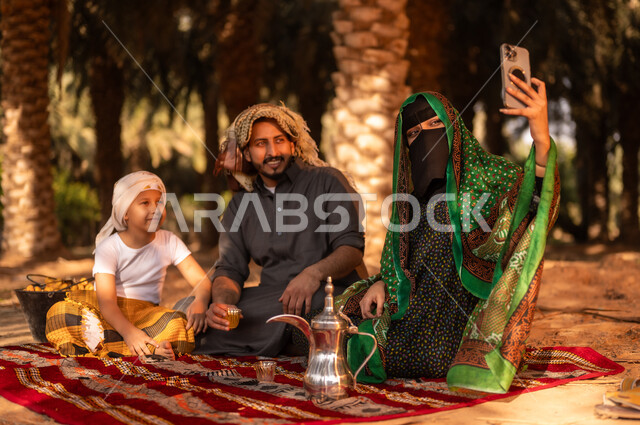 قضاء وقت الراحة في الهواء الطلق مع العائلة، فلاح عربي خليجي سعودي مبتسم يرتدي الثوب ويضع الشماغ على رأسه للحماية من الشمس  ويجلس على الأرض مع زوجته وولده في المزرعة، أخذ صور السيلفي للذكرى بعد العمل في مزارع النخيل المثمرة، توريث المهن الزراعية التقليدية