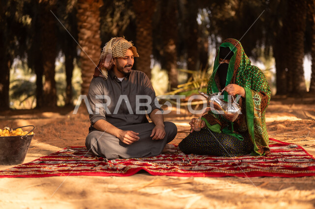أخذ قسط من الراحة وتبادل الأحاديث بعد العمل في مزارع النخيل المثمرة، قضاء وقت الراحة في الهواء الطلق، فلاح عربي خليجي سعودي مبتسم يرتدي الثوب ويضع الشماغ على رأسه للحماية من الشمس ويجلس على الأرض مع زوجته في المزرعة،  مهن زراعية تقليدية، ايماءات السرور والبهجة