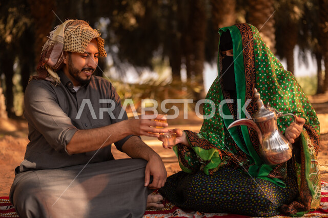 قضاء وقت الراحة في الهواء الطلق، فلاح عربي خليجي سعودي مبتسم يرتدي الثوب ويضع الشماغ على رأسه للحماية من الشمس ويجلس على الأرض مع زوجته في المزرعة، أخذ قسط من الراحة وتبادل الأحاديث بعد العمل في مزارع النخيل المثمرة، مهن زراعية تقليدية، ايماءات السرور والبهجة