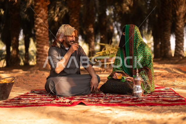 ايماءات السرور والبهجة، أخذ قسط من الراحة وتبادل الأحاديث بعد العمل في مزارع النخيل المثمرة، قضاء وقت الراحة في الهواء الطلق، فلاح عربي خليجي سعودي مبتسم يرتدي الثوب ويضع الشماغ على رأسه للحماية من الشمس ويجلس على الأرض مع زوجته في المزرعة، مهن زراعية تقليدية