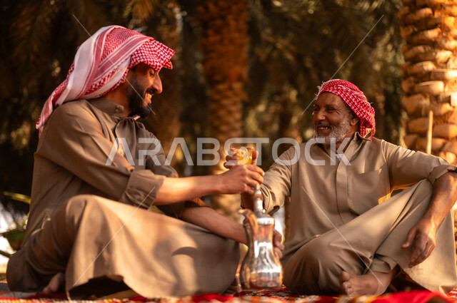 Taking a break and exchanging conversations after working in the fruitful palm farms, spending rest time in the open air, a smiling Saudi Arabian Gulf farmer wearing a thobe and wearing a shemagh on his head to protect from the sun, sitting on the ground with his father on the farm, traditional agricultural occupations