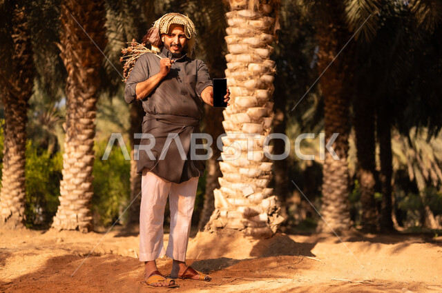 موسم الحصاد وجني التمور في المزرعة، قضاء وقت العمل في الهواء الطلق، محاصيل زراعية محلية سعودية، فلاح عربي خليجي سعودي مبتسم يرتدي الثوب والعمامة ينظر إلى الكاميرا ويحمل بيده الهاتف المحمول وعذق التمر، إيماءات السعادة والسرور، مهن زراعية تقليدية، عرض شاشة سوداء فارغة