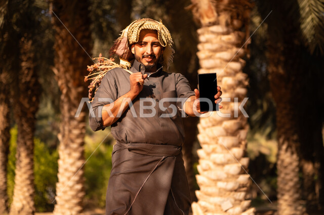 قضاء وقت العمل في الهواء الطلق، محاصيل زراعية محلية سعودية، موسم الحصاد وجني التمور في المزرعة، فلاح عربي خليجي سعودي مبتسم يرتدي الثوب والعمامة يحمل بيده الهاتف المحمول وعذق التمر، إيماءات السعادة والسرور، مهن زراعية تقليدية، عرض شاشة سوداء فارغة