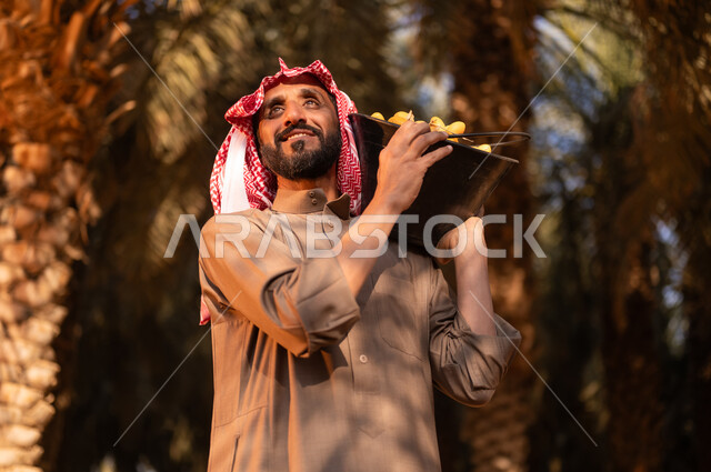 موسم الحصاد وجني التمور في المزرعة، قضاء وقت العمل في الهواء الطلق، فلاح عربي خليجي سعودي مبتسم يرتدي الثوب والعمامة ينظر إلى الأعلى ويحمل على كتفه أداة الحفر، الزراعة والاهتمام بالأشجار المثمرة في مزارع المملكة، محاصيل زراعية محلية، مهن زراعية تقيدية