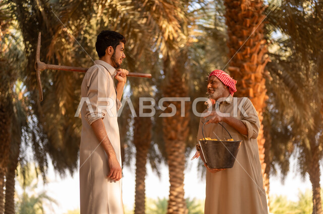 توارث مهنة الزراعة من الأجداد، فلاح عربي خليجي سعودي يرتدي الثوب والعمامة يحمل بيده البلح ويقوم بالتحدث مع ابنه في مزرعة النخيل، شاب يقوم بوضع أداة الحفر على كتفه في مزارع النخيل، موسم حصاد التمور في المملكة، محاصيل زراعية محلية، مهن تقليدية زراعية سعودية