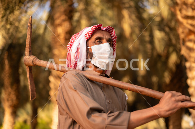 الزراعة والاهتمام بالأشجار المثمرة في مزارع المملكة، قضاء وقت العمل في الهواء الطلق، فلاح عربي خليجي سعودي يرتدي الثوب والعمامة ويضع الكمامة للحماية من الغبار ويحمل على كتفه أداة الحفر، استخدام أدوات شعبية قديمة في العمل في المزرعة، مهن تقليدية سعودية