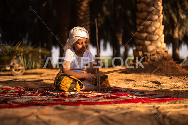 Enjoying leisure time with fun and entertainment on the Kingdom’s farms, preserving a rich agricultural heritage, spending time outdoors, a smiling Saudi Gulf Arab child wearing a traditional costume and a turban sitting on the ground and playing with wooden sticks inside a palm farm, playing in the old folk way