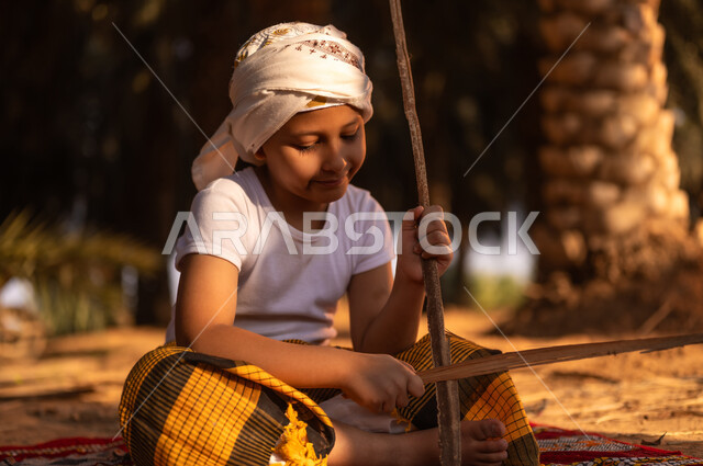 Enjoying leisure time with fun and entertainment on the Kingdom’s farms, spending time outdoors, a smiling Saudi Gulf Arab child wearing a traditional costume and a turban sitting on the ground and playing with wooden sticks inside a palm farm, playing in the old folk way