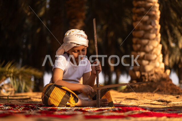 Spending time outdoors, a smiling Saudi Arabian Gulf child wearing a traditional costume and a turban, sitting on the ground and playing with wooden sticks inside a palm farm, playing in the old folk way, enjoying his leisure time with amusement and amusement in the Kingdom’s farms
