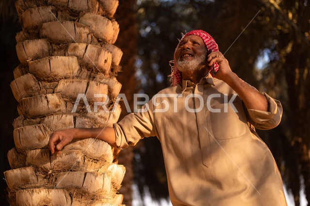 بيع وشراء التمر أونلاين، ايماءات البهجة والفرح أثناء قضاء وقت الاستراحة في الهواء الطلق، فلاح سعودي خليجي مبتسم يرتدي الثوب والعمامة ينظر إلى النخيل ويقوم باجراء مكالمة هاتفية من خلال الجوال ويستند على شجرة النخيل، مهن تقليدية سعودية