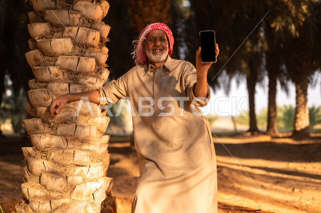ايماءات البهجة والفرح أثناء قضاء وقت الاستراحة في الهواء الطلق، فلاح سعودي خليجي مبتسم يرتدي الثوب والعمامة ينظر إلى الكاميرا ويحمل بيده الهاتف المحمول داخل مزرعة النخيل المثمرة بعد انجاز العمل، بيع وشراء التمر أونلاين، مهن تقليدية سعودية، عرض شاشة سوداء فارغة