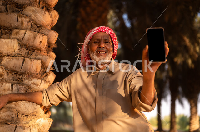 قضاء وقت الاستراحة في الهواء الطلق، فلاح سعودي خليجي مبتسم يرتدي الثوب والعمامة ينظر إلى الكاميرا ويحمل بيده الهاتف المحمول داخل مزرعة النخيل المثمرة بعد انجاز العمل، بيع وشراء التمر أونلاين، ايماءات البهجة والفرح، مهن تقليدية سعودية، عرض شاشة سوداء فارغة