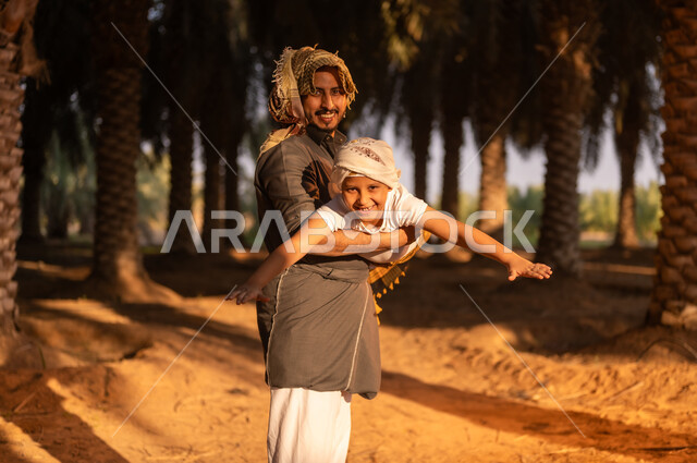 الحفاظ على تراث زراعي غني، قضاء وقت الراحة في الهواء الطلق، فلاح عربي خليجي سعودي مبتسم يرتدي زي الحصاد والعمامة يقوم بحمل ابنه واللعب معه داخل مزارع النخيل المثمرة، ايماءات الفرح والسرور، مهن تقليدية سعودية، محاصيل زراعية محلية