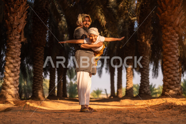 قضاء وقت الراحة في الهواء الطلق، فلاح عربي خليجي سعودي مبتسم يرتدي زي الحصاد والعمامة يقوم بحمل ابنه واللعب معه داخل مزارع النخيل المثمرة، الحفاظ على تراث زراعي غني، ايماءات الفرح والسرور، مهن تقليدية سعودية، محاصيل زراعية محلية