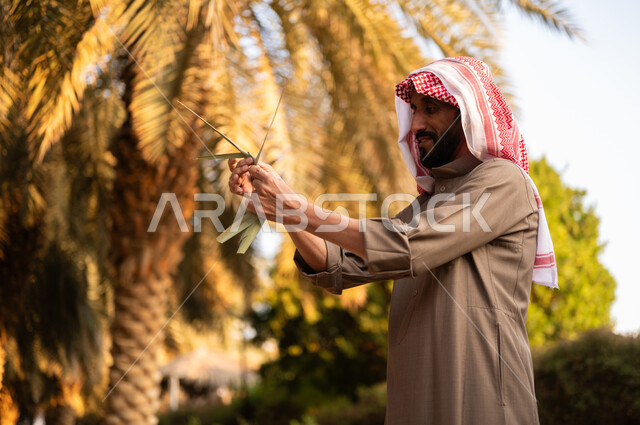 استخدام أدوات شعبية قديمة للعمل في المزرعة، حرفة يدوية شعبية تقليدية، نسج سعف النخيل الأخضر، أوراق النخل المنسوجة يدويا، قضاء وقت العمل في الهواء الطلق، فلاح عربي خليجي سعودي مبتسم يرتدي الثوب والعمامة يقوم بصناعة شيء ما من الخوص، مهن تقليدية سعودية