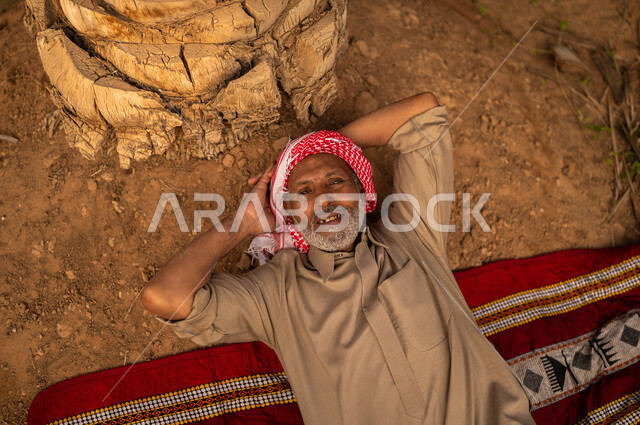 قضاء وقت الاستراحة في الهواء الطلق، فلاح عربي خليجي سعودي مبتسم يرتدي الثوب والعمامة يستلقي على بساط بعد العمل في المزرعة ينظر إلى الكاميرا بسعادة، ايماءات الاستمتاع بالاسترخاء على تربة الأرض السعودية، مهن تقليدية سعودية