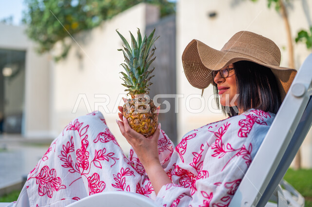 الاستجمام والاسترخاء في شاليهات المملكة العربية السعودية، صورة من الخلف لامرأة عربية خليجية سعودية ترتدي قبعة قش و تجلس على كرسي مريح أمام المسبح تحمل بيدها حبة أناناس، الفواكه الصيفية الطازجة، الاستمتاع بعطلة سعيدة