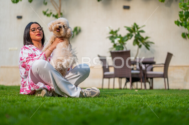 الشعور بالسعادة أثناء قضاء وقت ممتع في أيام العطلة، اللعب والمرح مع الحيوانات الأليفة، مفهوم الرفق بالحيوان، تربية واقتناء الكلاب المنزلية، امرأة عربية خليجية سعودية تجلس على العشب وتحمل جرو صغير